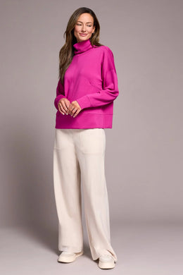 Woman wearing a bright pink funnel neck knit top, white pants and white sneakers against a plain background.