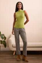 Load image into Gallery viewer, Woman wearing a green sleeveless top, gray pants and brown boots standing in a room with a beige wall and a plant.
