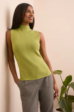Woman wearing a green sleeveless top and gray pants against a beige wall with a plant.