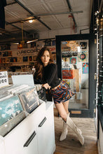 Load image into Gallery viewer, Woman in a record store with vinyl records and shelves in the background, wearing a black sweater, abstract print mini skort and tall white boots.
