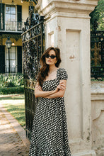 Load image into Gallery viewer, Woman in a brown and white polka dot dress and sunglasses standing next to a stone column in an elegant outdoor setting.
