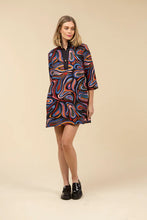 Load image into Gallery viewer, Woman wearing a colorful abstract patterned dress and black loafers against a light beige background.
