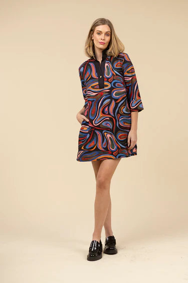 Woman wearing a colorful abstract patterned dress and black loafers against a light beige background.