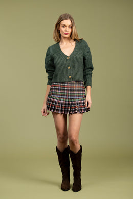 Woman wearing a green cardigan,  multi-color plaid skirt and brown boots against a pale green background.