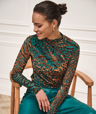 Woman wearing an animal print top in burnout velvet in teal and brown and teal pants sitting on a wooden chair.