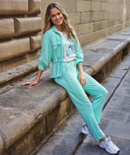 Load image into Gallery viewer, Woman in a mint green tracksuit and colorful tee and tennis shoes sitting on stone steps with a stone wall background.
