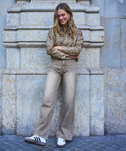 Load image into Gallery viewer, Woman wearing an animal print jacket, light caramel faded jean and white sneakers leaning against a stone wall.
