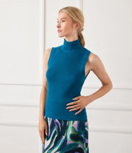 Load image into Gallery viewer, Woman wearing a teal blue sleeveless ribbed turtleneck knit top and colorful skirt against a white wall.
