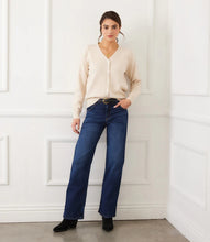 Load image into Gallery viewer, Woman wearing a beige cardigan, blue jeans, heart necklace, black belt and black boots against a white wall.
