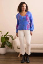 Load image into Gallery viewer, Woman wearing a blue sweater with colorful patterns, off-white pants and black boots standing in front of a beige chair and a plant.
