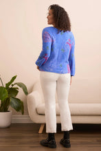 Load image into Gallery viewer, Woman wearing a blue sweater with colorful patterns, off-white pants and black boots standing in front of a beige chair and a plant.
