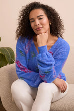 Load image into Gallery viewer, Woman wearing a blue sweater with colorful patterns and off-white pants sitting on a beige chair.
