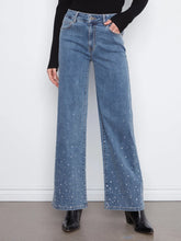 Load image into Gallery viewer, A model stands against a white wall wearing a black ribbed long-sleeve mock-neck top and high-waisted, wide-leg blue jeans with rhinestone embellishments near the lower hems, paired with black ankle boots.
