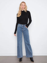 Load image into Gallery viewer, A woman stands against a white wall wearing a black ribbed long-sleeve mock-neck top and high-waisted, wide-leg blue jeans with rhinestone embellishments near the lower hems, paired with black ankle boots.
