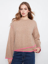 Load image into Gallery viewer, A woman wearing a cropped, long-sleeve knit sweater in a heathered tan-beige color with bright pink contrast trim along the collar, cuffs, and hem, paired with dark blue jeans against a plain white studio background.
