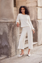 Load image into Gallery viewer, Woman wearing a sparkle blouse with white crop pants and white sandals standing against a stone building.
