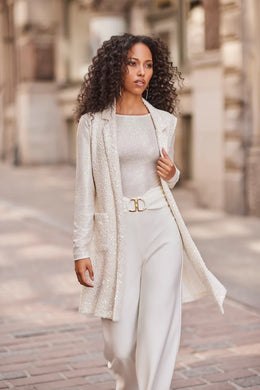 Woman wearing white sparkle long sleeve top, white sparkle long vest and white pants walking on a brick path with a stone building in the background.